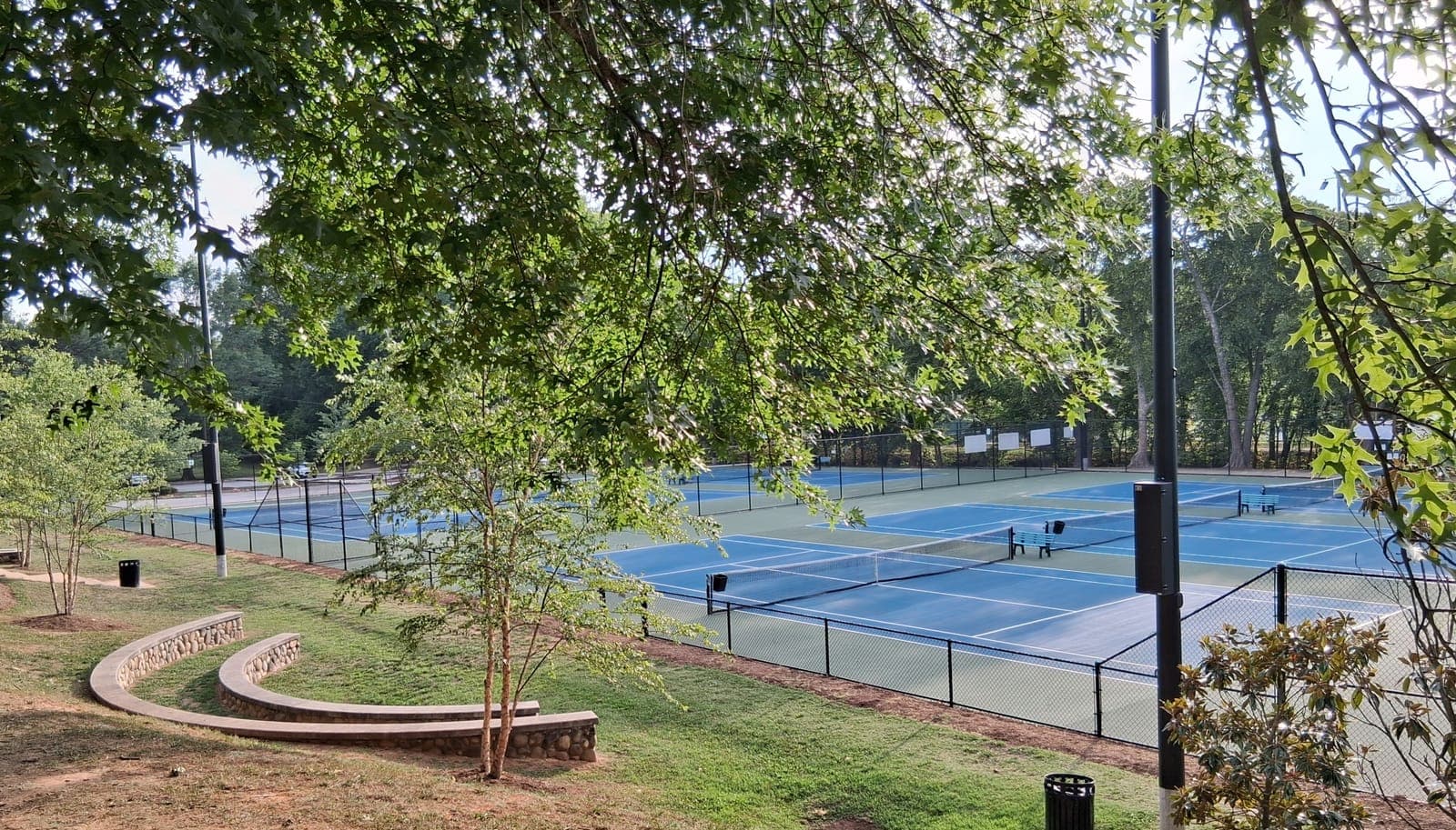 Empty blue hard-surface tennis courts under shade trees on a quiet morning, for an AP Lang argument essay on Naomi Osaka and living in the present moment