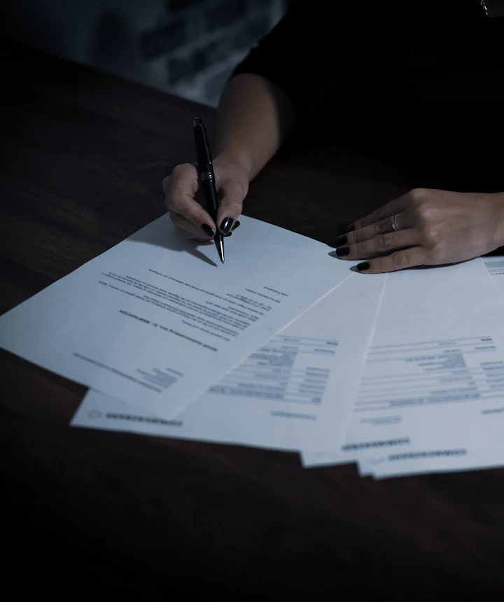 A hand holding a pen over a spread of papers on a dark desk